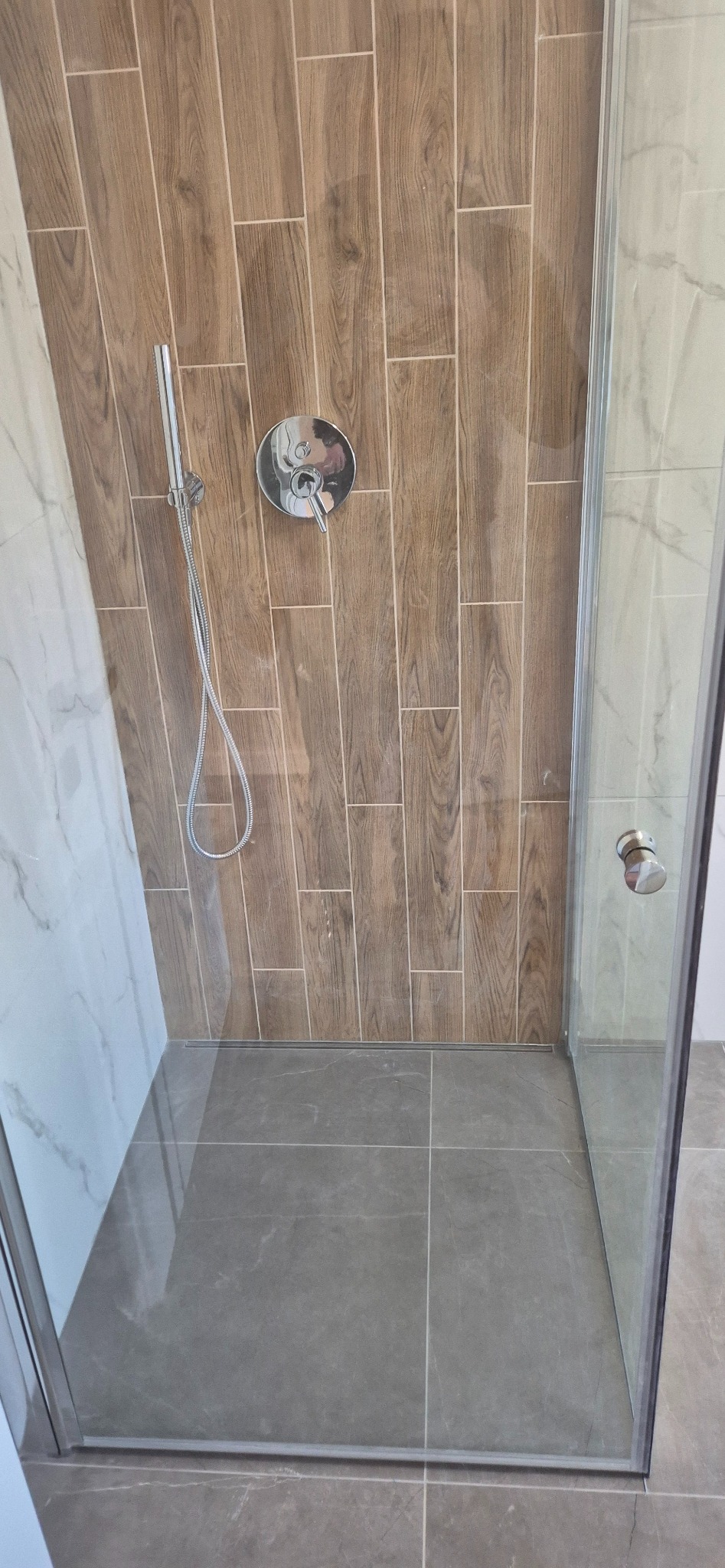 Kabina prysznicowa z drewnopodobnymi płytkami na ścianie i szarą podłogą. Widoczna bateria i słuchawka prysznicowa. Minimalistyczny design.