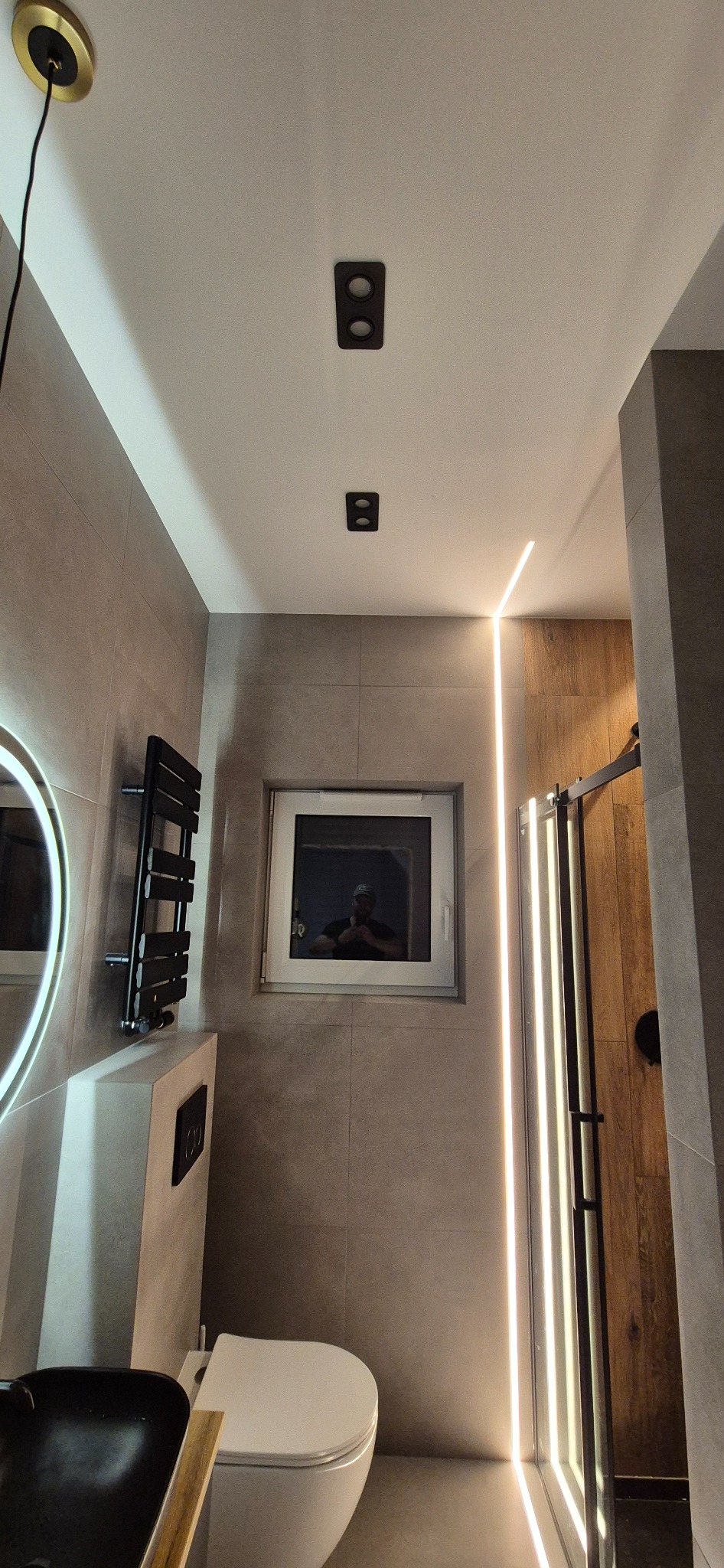 Nowoczesna łazienka z szarymi płytkami, podświetlanym prysznicem i okrągłym lustrem z podświetleniem. Widoczny czarny grzejnik drabinkowy i toaleta.