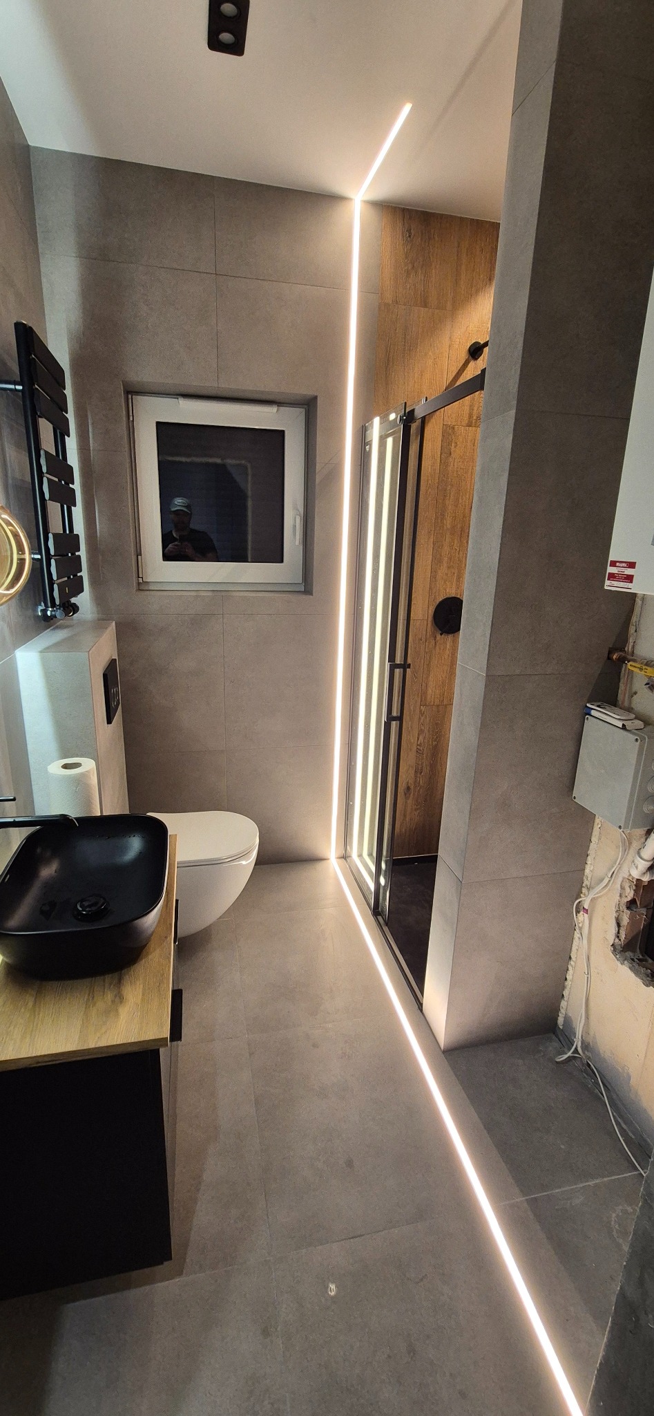 Nowoczesna łazienka z szarą glazurą, czarną umywalką nablatową i liniowym oświetleniem LED wzdłuż kabiny prysznicowej. Widoczne niedokończone prace przy instalacji.