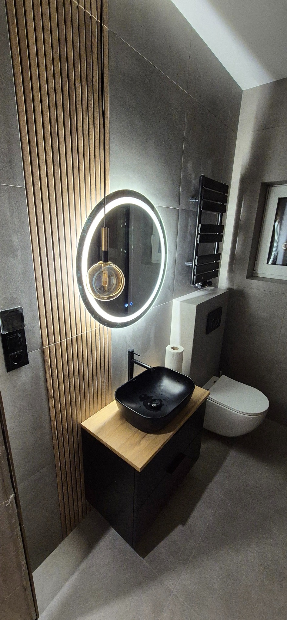 Nowoczesna łazienka z drewnianymi lamelami, okrągłym lustrem z podświetleniem LED i czarną umywalką nablatową. Szare płytki i minimalistyczny design.