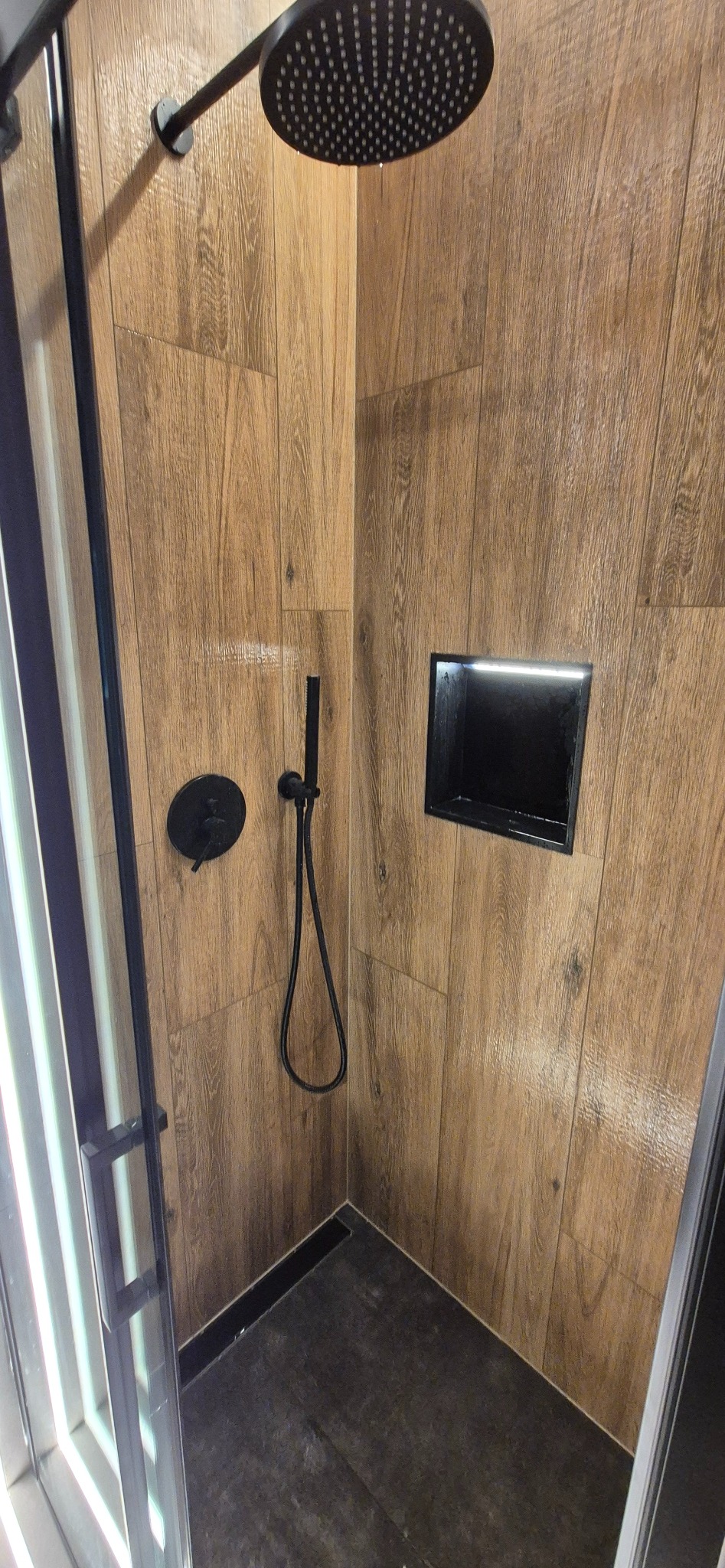Nowoczesna kabina prysznicowa z drewnopodobnymi płytkami, czarną armaturą i wnęką z podświetleniem LED. Minimalistyczny design, eleganckie wykończenie.