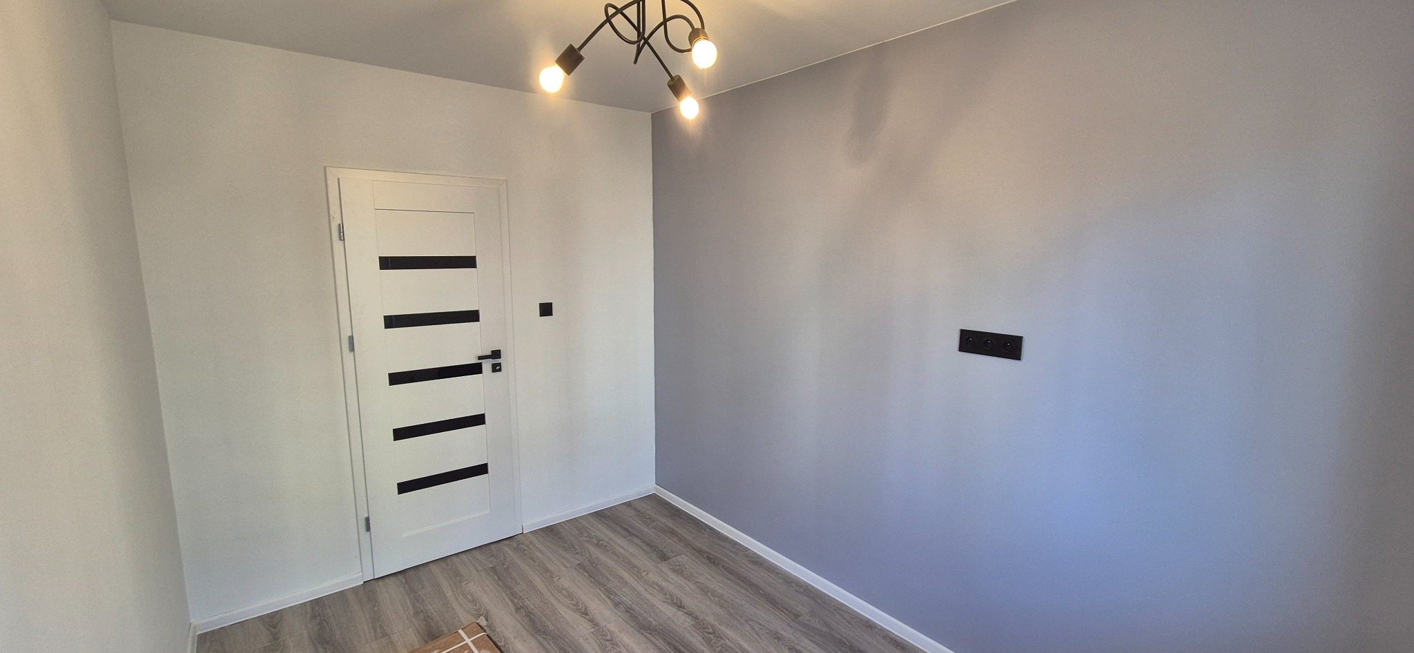 Pokój po remoncie: szare ściany, drewnopodobna podłoga, białe drzwi z czarnymi paskami i nowoczesna lampa sufitowa. Widoczne gniazdka elektryczne.