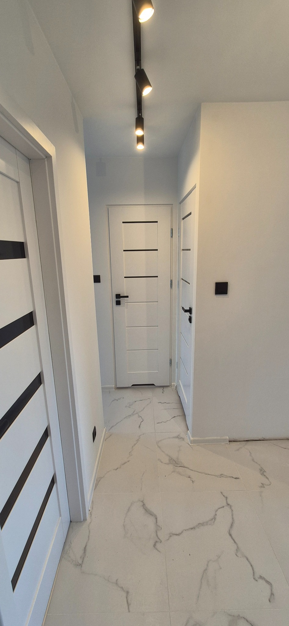 Białe wnętrze korytarza z marmurową podłogą, oświetleniem punktowym i trzema parami białych drzwi z czarnymi akcentami. Minimalistyczny design.