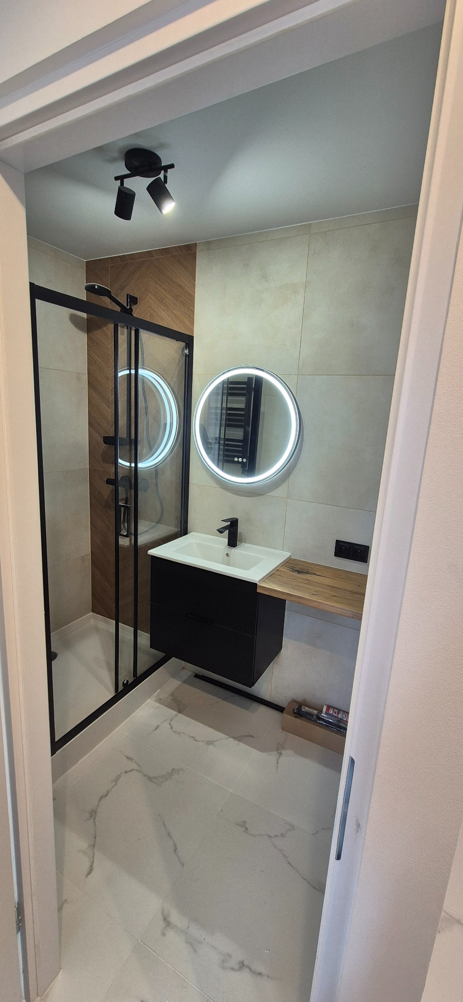 Nowoczesna łazienka po remoncie: kabina prysznicowa typu walk-in z czarną ramą, okrągłe lustro z podświetleniem LED i czarna szafka pod umywalką z drewnianym blatem.