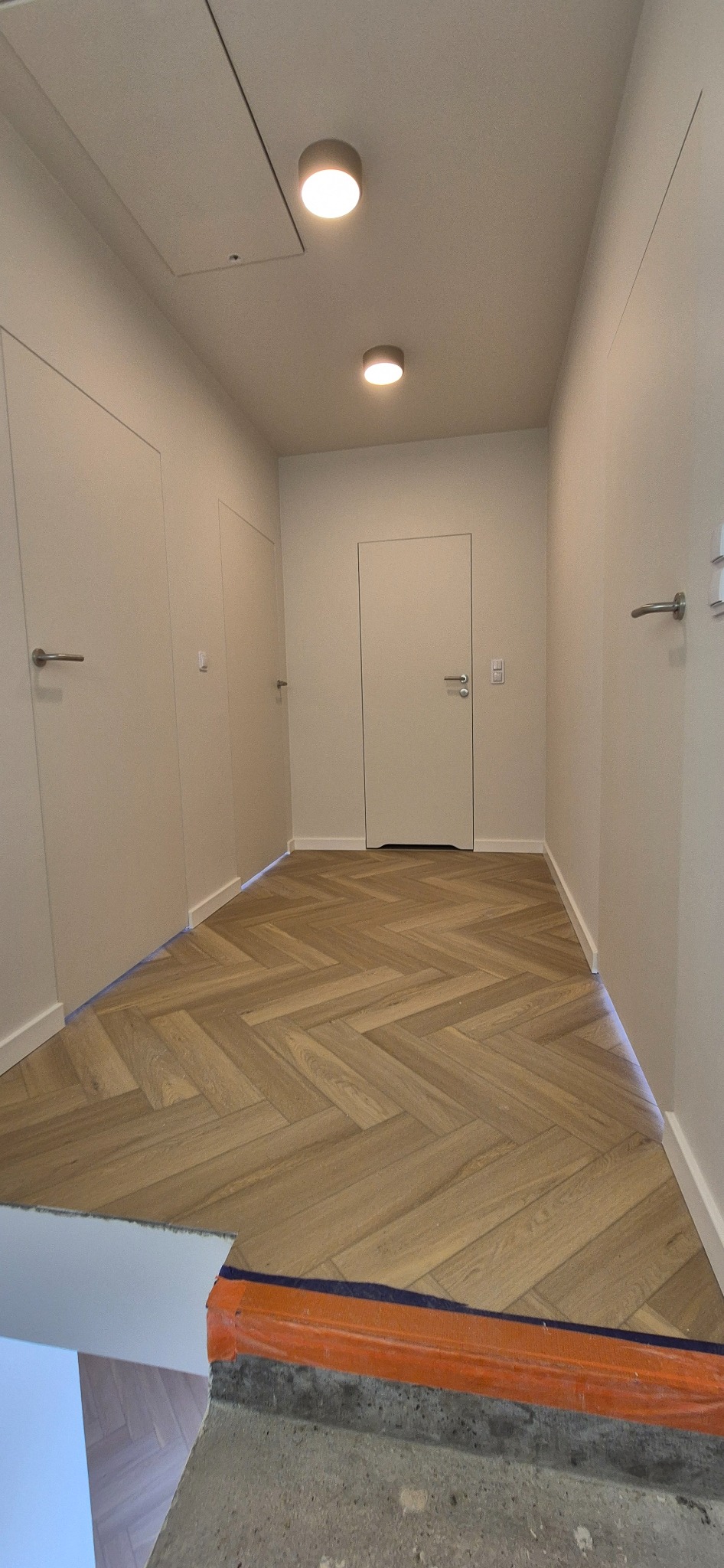 Korytarz z parkietem ułożonym w jodełkę. Widoczne drzwi w kolorze ścian i oświetlenie sufitowe. Fragment posadzki przed wykończeniem.