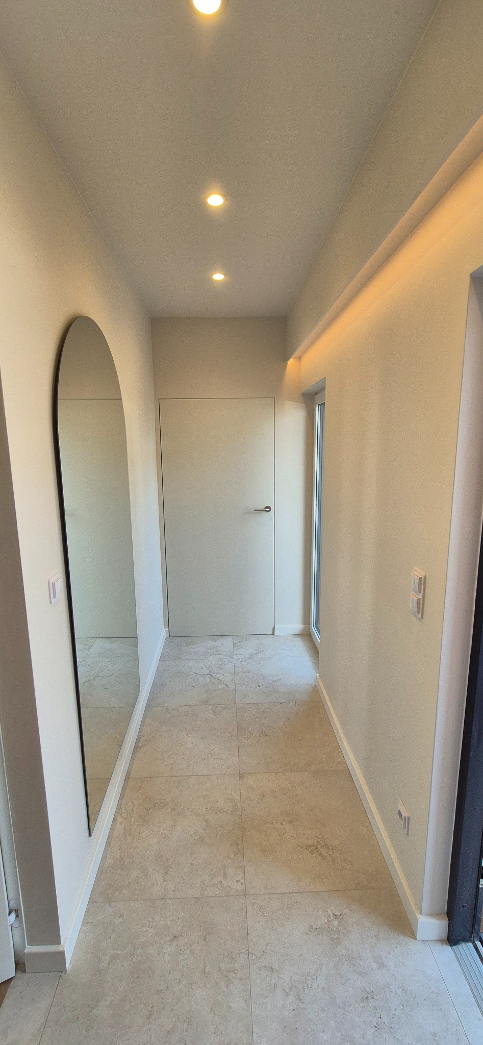 Jasny korytarz z beżowymi płytkami podłogowymi i minimalistycznym lustrem w kształcie łuku. Sufit z oświetleniem punktowym i ukrytym paskiem LED.