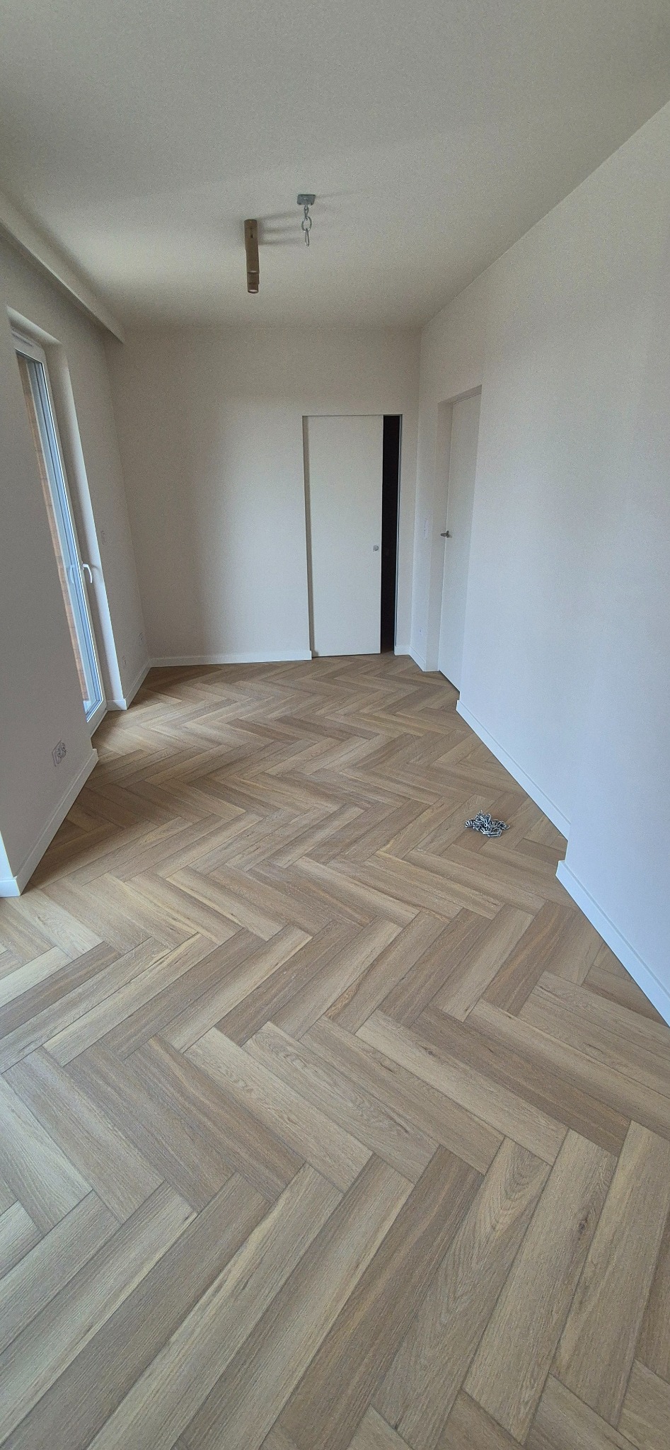 Jasne panele ułożone w jodełkę francuską w pustym pokoju z białymi ścianami. Widoczne okno, drzwi przesuwne i elementy instalacji oświetleniowej na suficie.