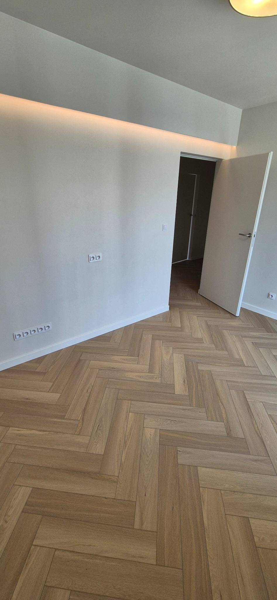 Jasne panele ułożone w jodełkę francuską, świeżo po montażu. Białe ściany z gniazdkami, otwarte drzwi do kolejnego pomieszczenia. Nowoczesne wykończenie wnętrza.