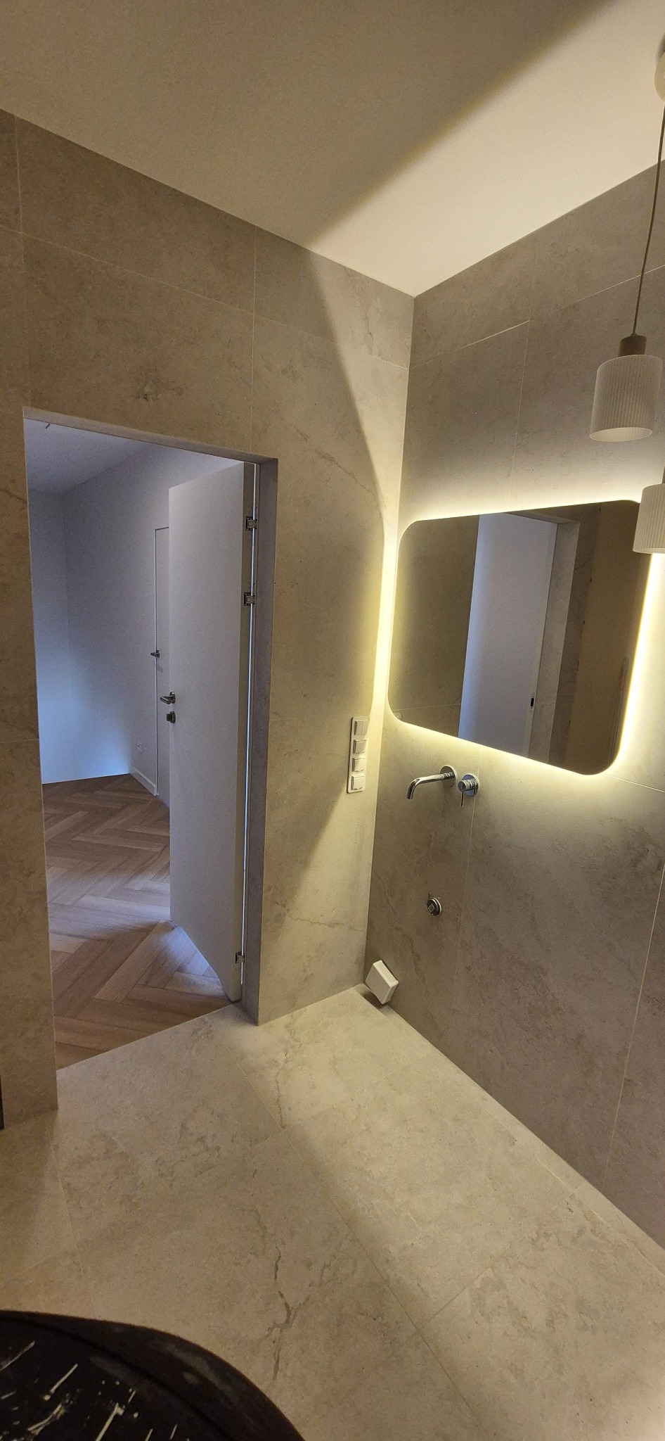 Beżowa łazienka z podświetlanym lustrem i otwartymi drzwiami, przez które widać parkiet ułożony w jodełkę. Minimalistyczny design, ciepłe oświetlenie.