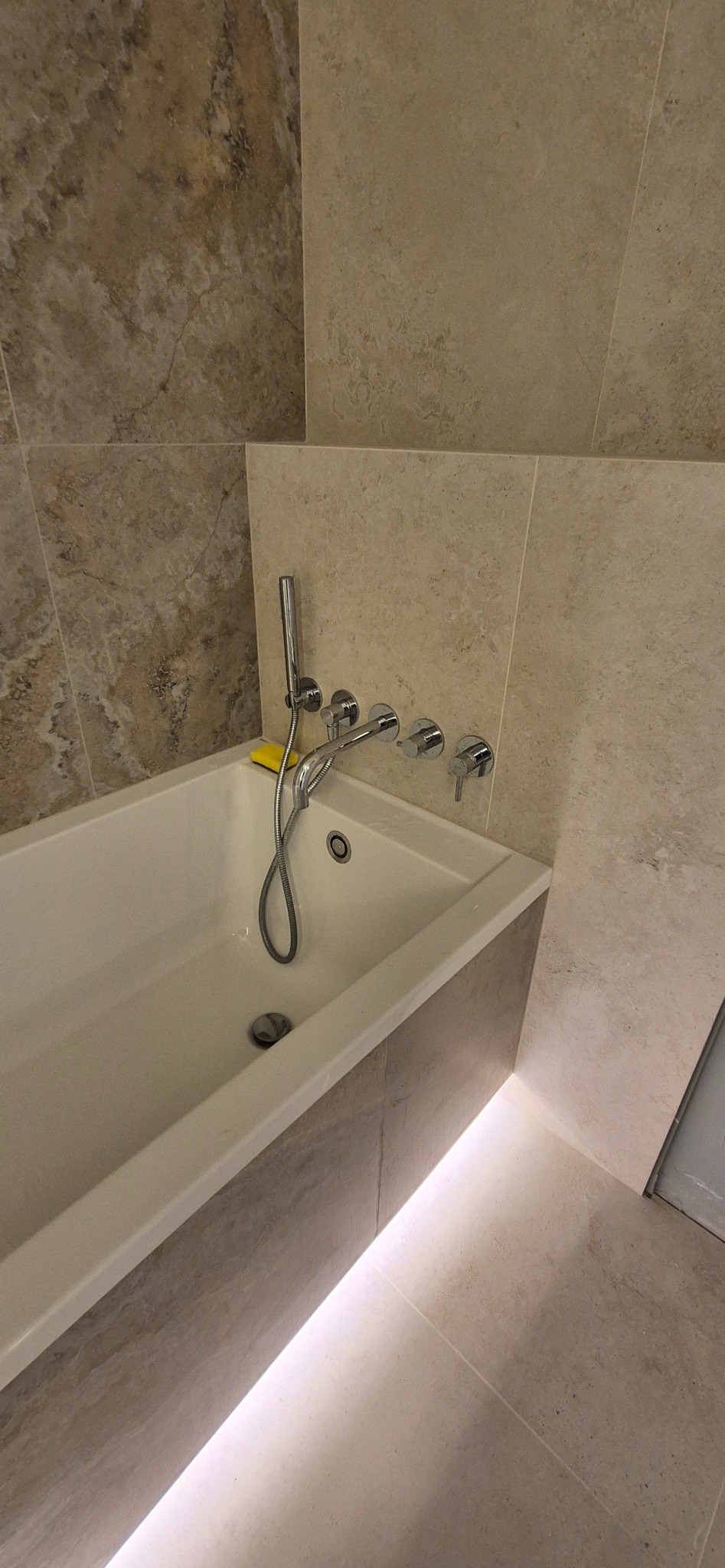 Wykończona łazienka: biała wanna z chromowaną armaturą i żółtą gąbką, obudowana płytkami w odcieniach beżu i brązu, z subtelnym podświetleniem LED u dołu.