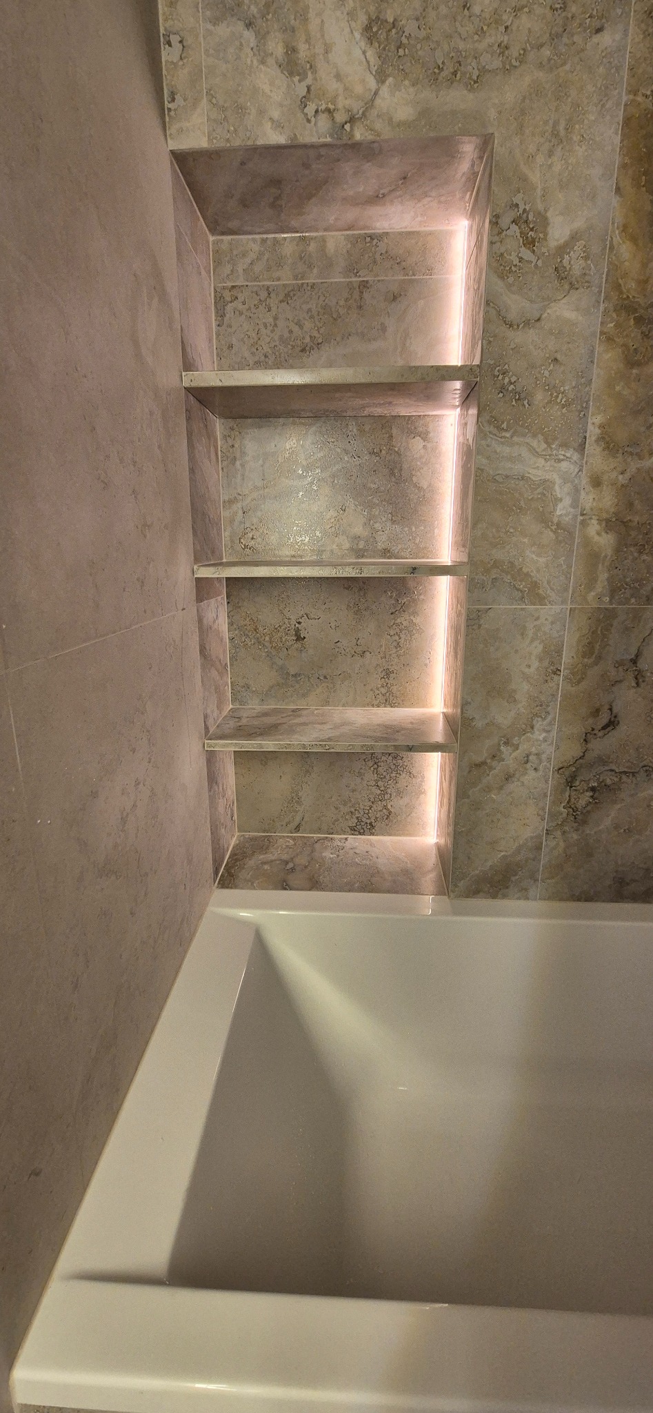 Wykończona wnęka z półkami podświetlonymi LED w łazience w Łodzi, obłożona płytkami imitującymi marmur, nad wanną. Detal wykończenia wnętrza.