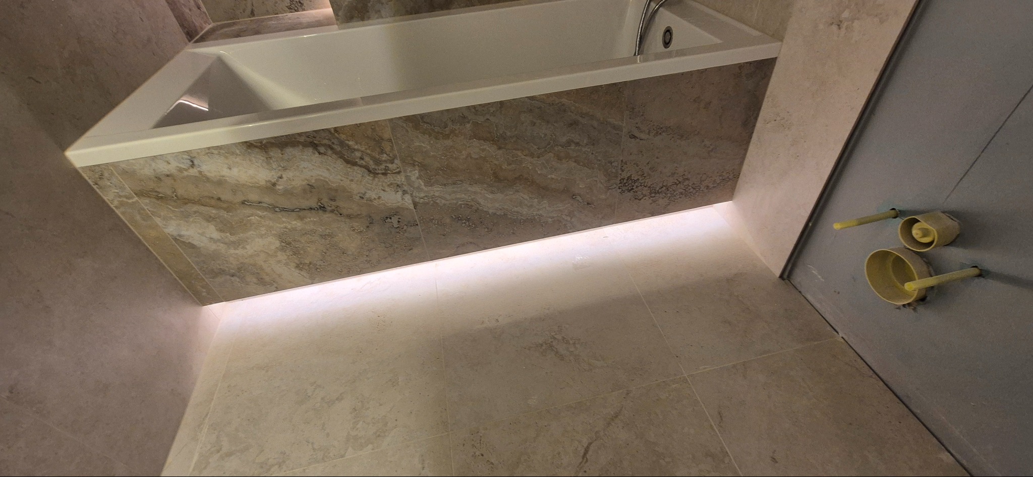 Wykończona łazienka z wanną obudowaną płytkami imitującymi marmur i podświetleniem LED. Widoczne przyłącza wodne na ścianie. Ukośne ujęcie.