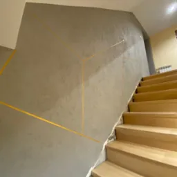 Ściana klatki schodowej z szarą fakturą i geometrycznymi, złotymi liniami, jasne drewniane schody.