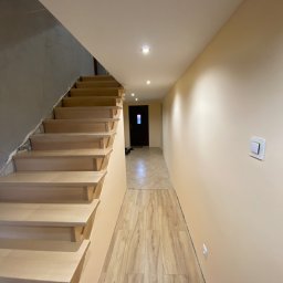 Paweł Tkaczyk - Korytarz w trakcie prac wykończeniowych: drewniane schody w jasnym odcieniu, ściana częściowo pomalowana na beżowo, a częściowo w stanie surowym, podłoga wyłożona panelami imitującymi drewno...