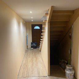 Paweł Tkaczyk - Korytarz w trakcie prac wykończeniowych: świeżo pomalowane ściany w odcieniach beżu, drewniane schody, częściowo ułożona podłoga z paneli, widoczne narzędzia i puszki z farbą.