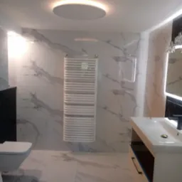 Nowoczesna łazienka z białymi płytkami imitującymi marmur, podwieszana toaleta, grzejnik drabinkowy i okrągłe oświetlenie sufitowe.