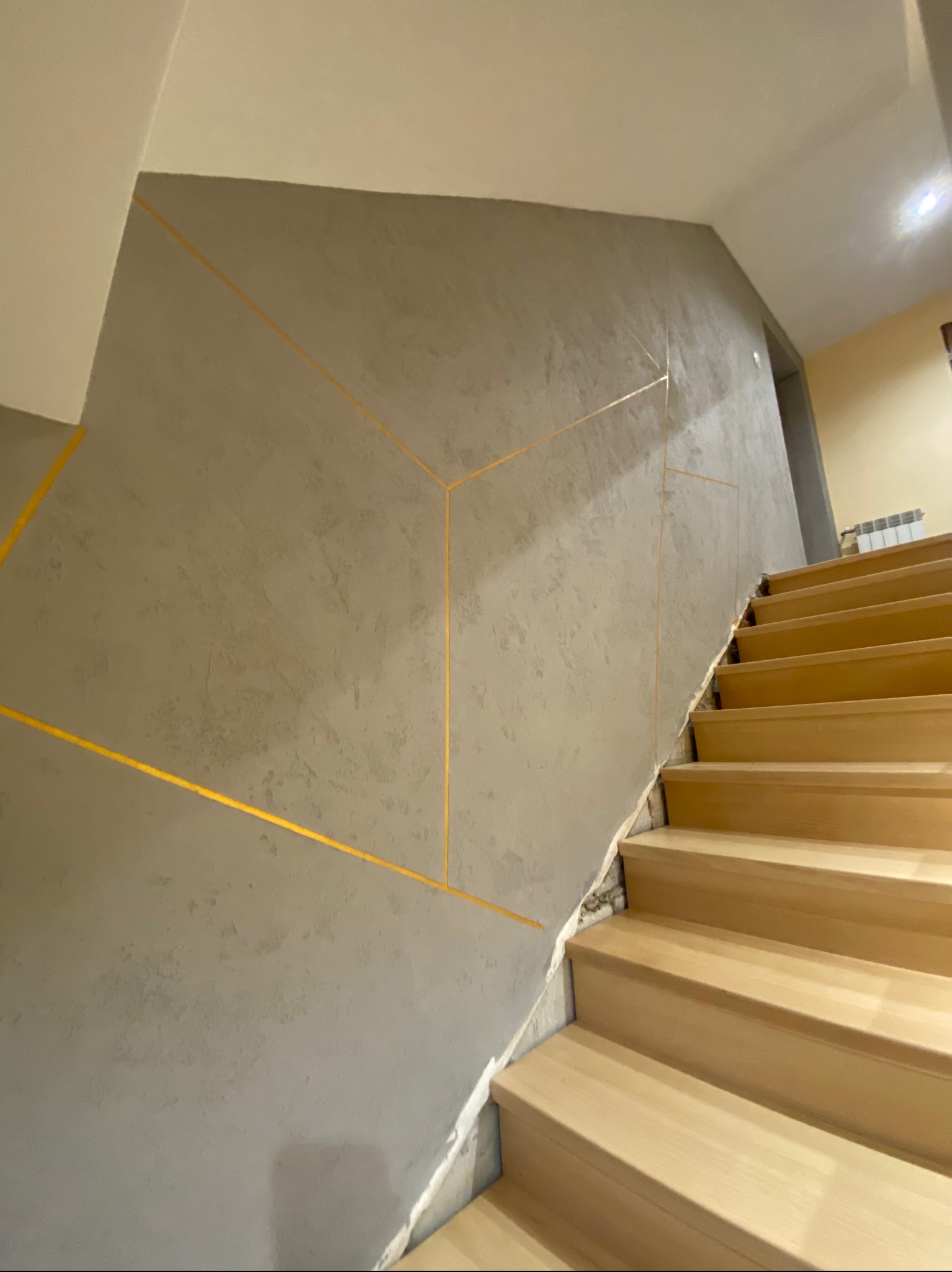 Ściana klatki schodowej z szarą fakturą i geometrycznymi, złotymi liniami, jasne drewniane schody.
