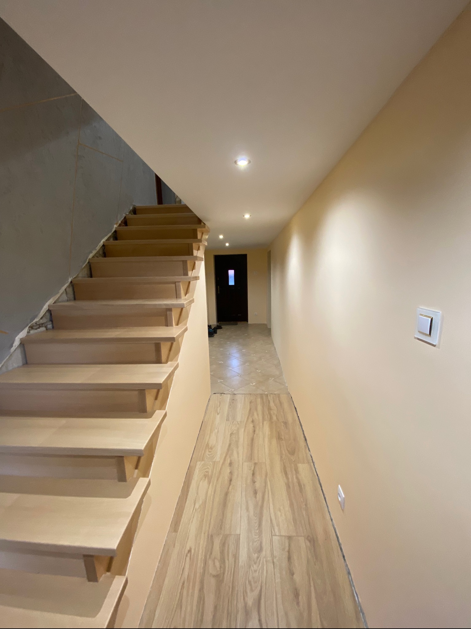 Korytarz w trakcie prac wykończeniowych: drewniane schody w jasnym odcieniu, ściana częściowo pomalowana na beżowo, a częściowo w stanie surowym, podłoga wyłożona panelami imitującymi drewno...