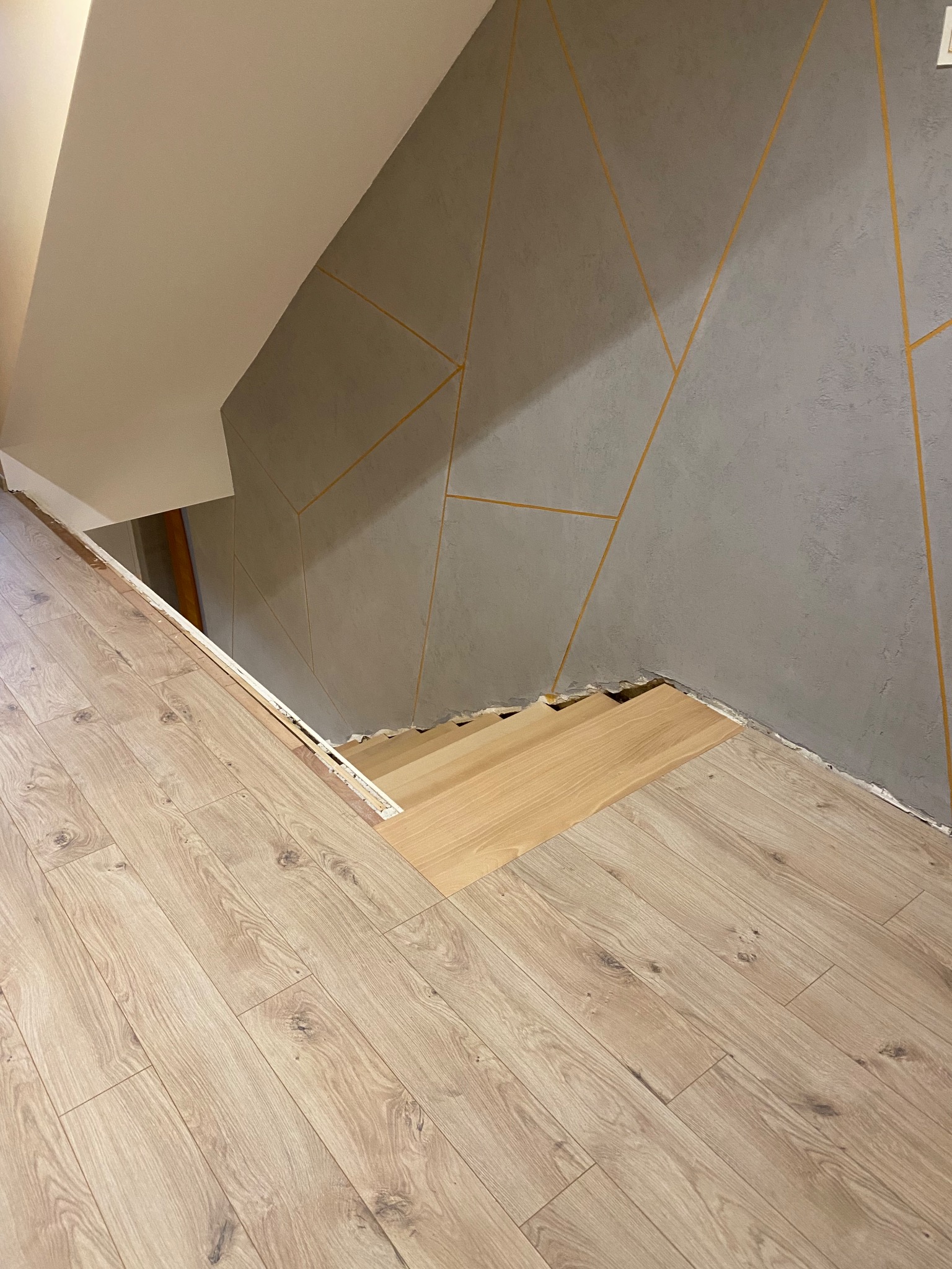 Fragment podłogi z jasnych paneli, częściowo ułożonych, z widocznym niedokończonym fragmentem przy schodach i szarą ścianą z geometrycznym wzorem w złotych liniach.