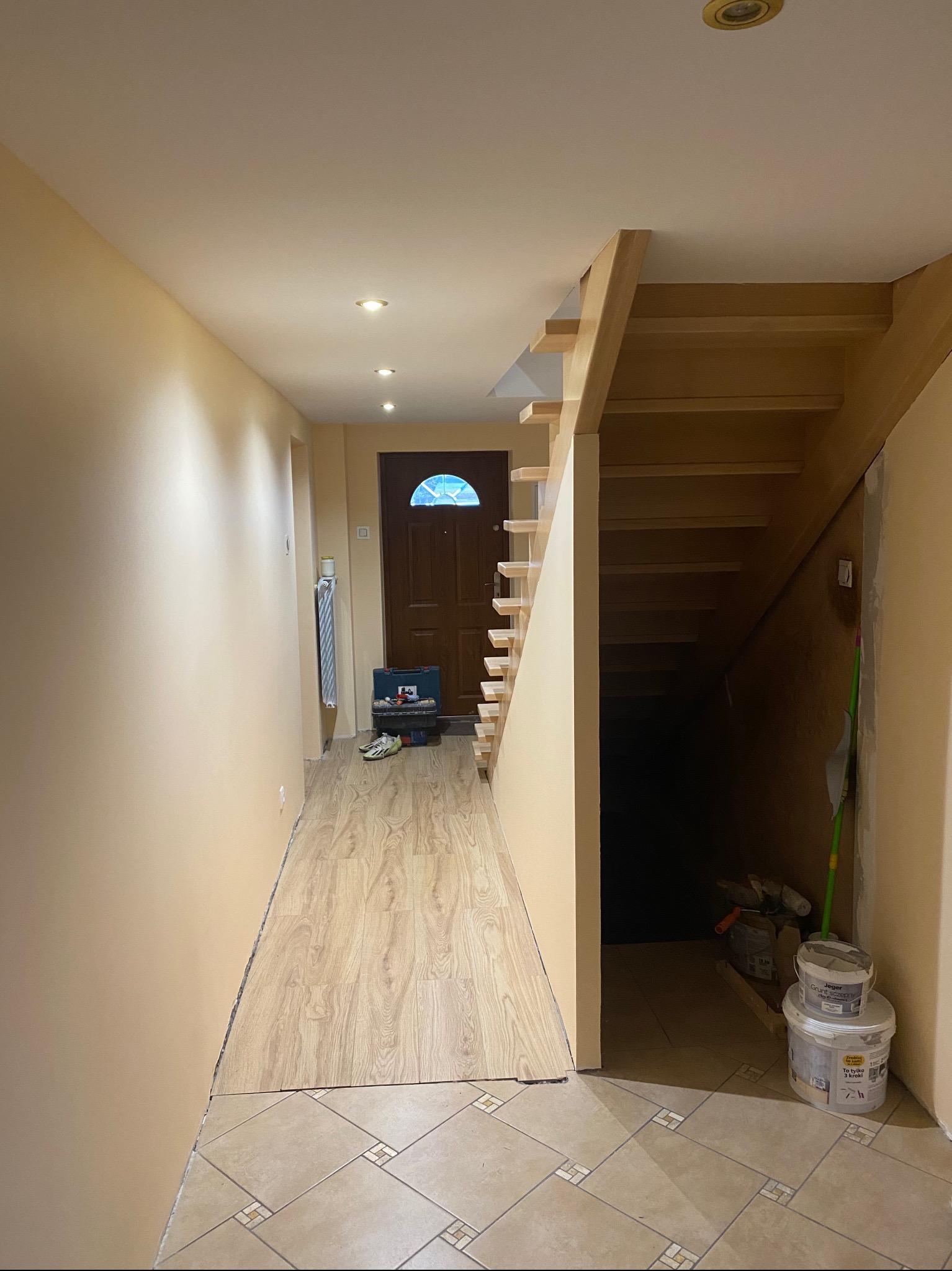 Korytarz w trakcie prac wykończeniowych: świeżo pomalowane ściany w odcieniach beżu, drewniane schody, częściowo ułożona podłoga z paneli, widoczne narzędzia i puszki z farbą.
