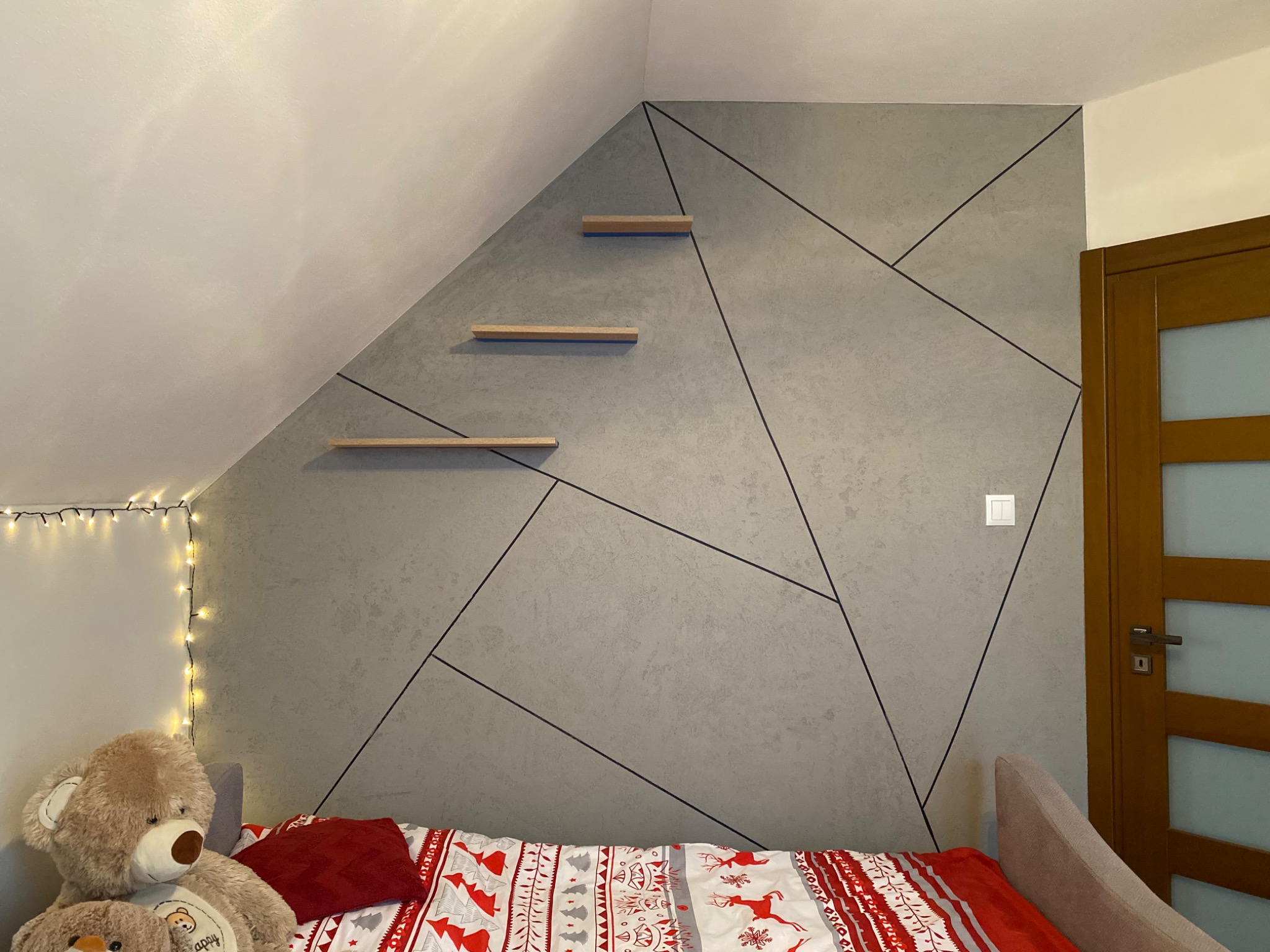 Ściana w pokoju na poddaszu z dekoracyjną szarą fakturą, czarnymi liniami tworzącymi geometryczny wzór i trzema drewnianymi półkami, nad łóżkiem z pościelą w świąteczne wzory i pluszowym misiem.