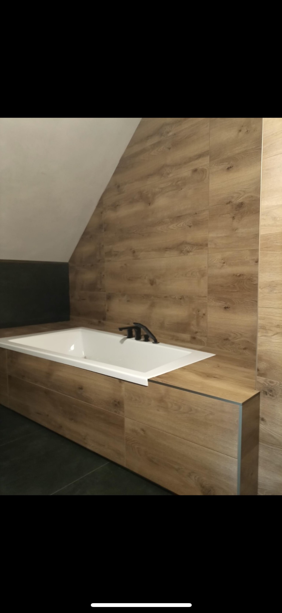 Nowoczesna łazienka z obudowaną wanną w drewnopodobnych płytkach, czarna bateria wannowa, ciemna podłoga, skośny sufit.