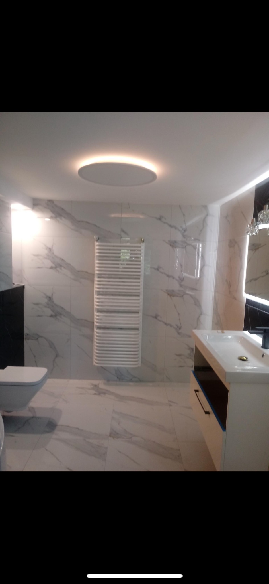 Nowoczesna łazienka z białymi płytkami imitującymi marmur, podwieszana toaleta, grzejnik drabinkowy i okrągłe oświetlenie sufitowe.