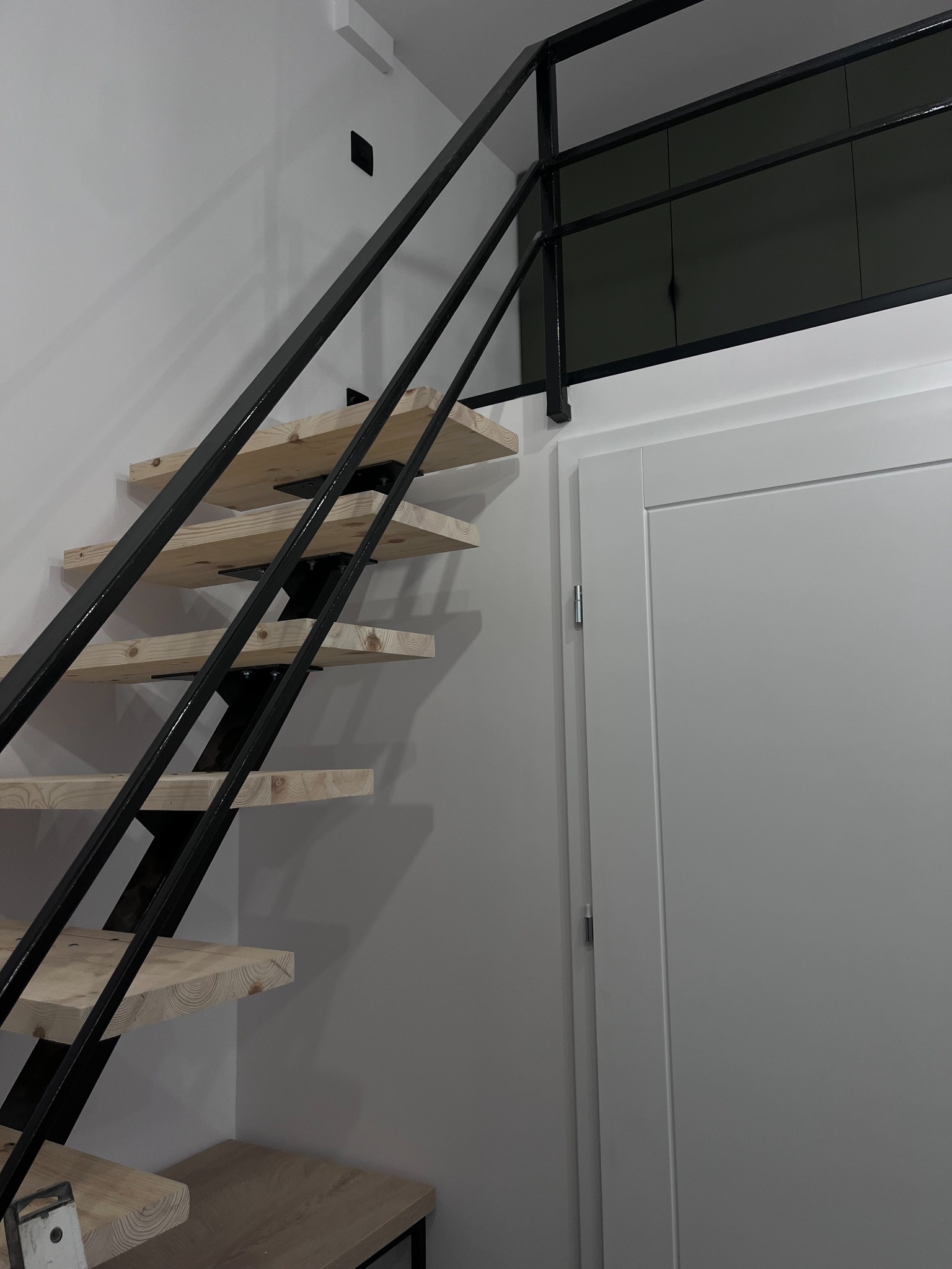 Nowoczesne schody ażurowe z drewnianymi stopniami i czarną metalową konstrukcją na tle białej ściany i drzwi.