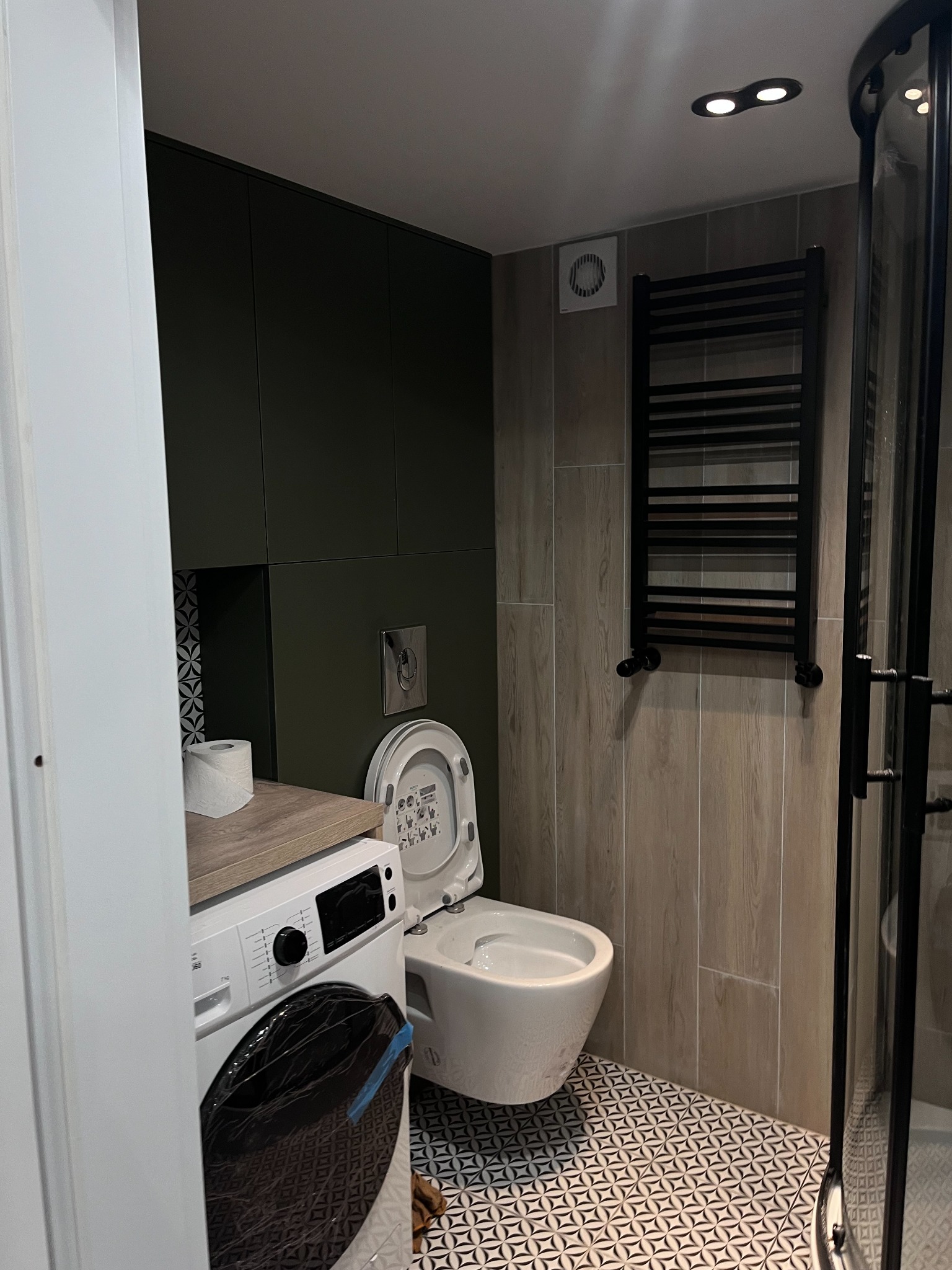 Nowoczesna łazienka z toaletą podwieszaną, pralką pod blatem, grzejnikiem drabinkowym i kabiną prysznicową z czarnymi profilami na podłodze w geometryczny wzór.