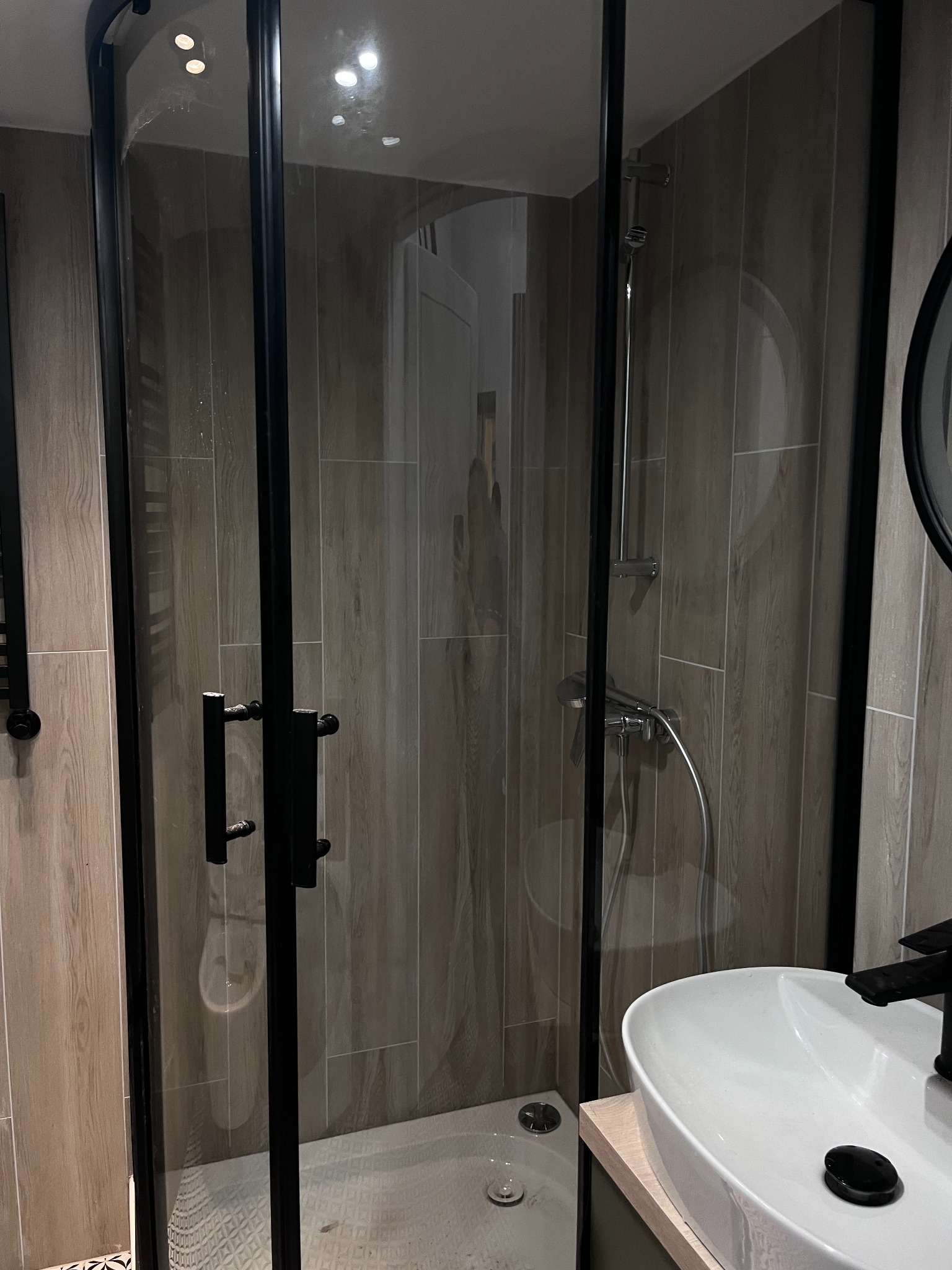 Nowoczesna kabina prysznicowa z czarnymi profilami i szklanymi drzwiami, widoczna umywalka z czarną armaturą, ściany wyłożone płytkami imitującymi drewno, wzorzysta podłoga.