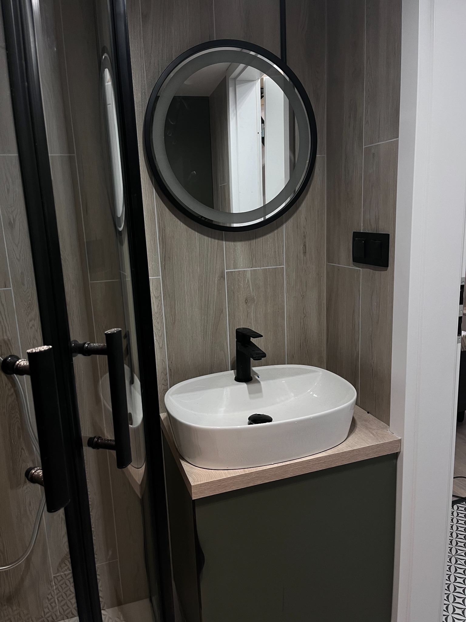 Nowoczesna łazienka z owalną białą umywalką na szafce w kolorze oliwkowym, czarną armaturą i okrągłym lustrem z podświetleniem LED, widoczna kabina prysznicowa z czarnymi profilami.