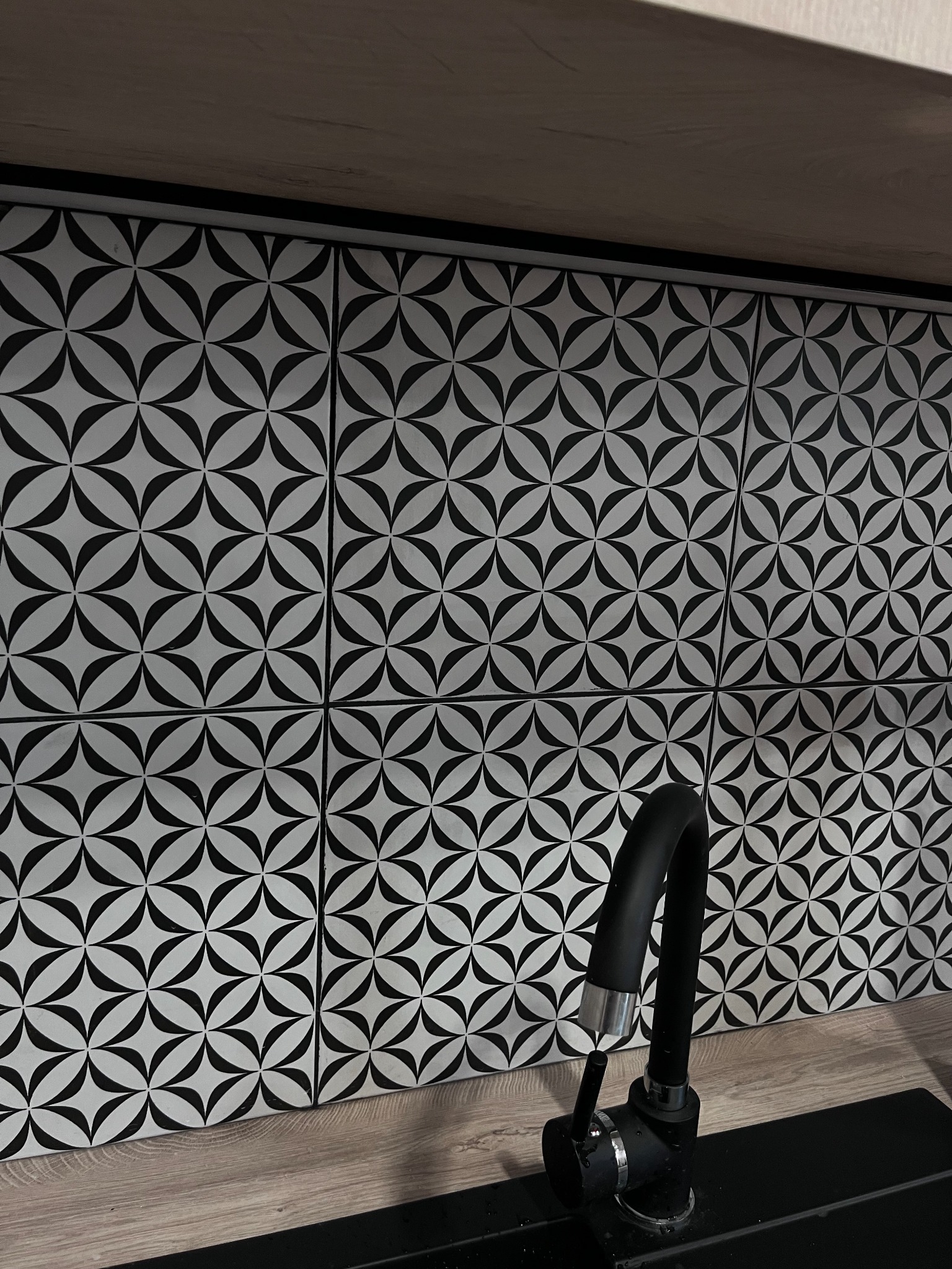 Ściana nad blatem kuchennym wyłożona białymi płytkami z czarnym geometrycznym wzorem kwiatowym, widoczny czarny kran z elementami chromowanymi.