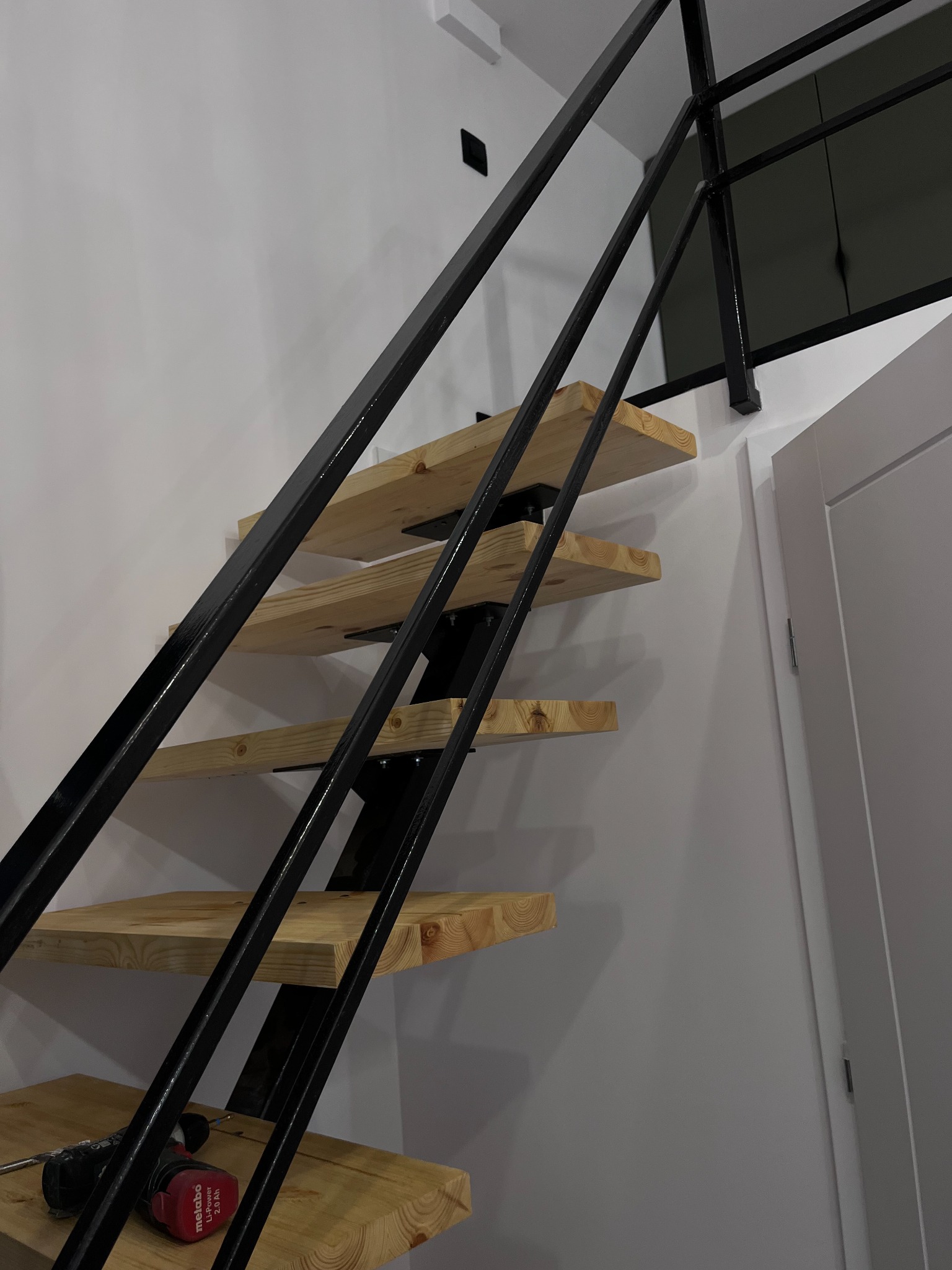 Metalowe schody z drewnianymi stopniami, widok z dołu, w tle biała ściana i drzwi, na jednym ze stopni wkrętarka akumulatorowa.