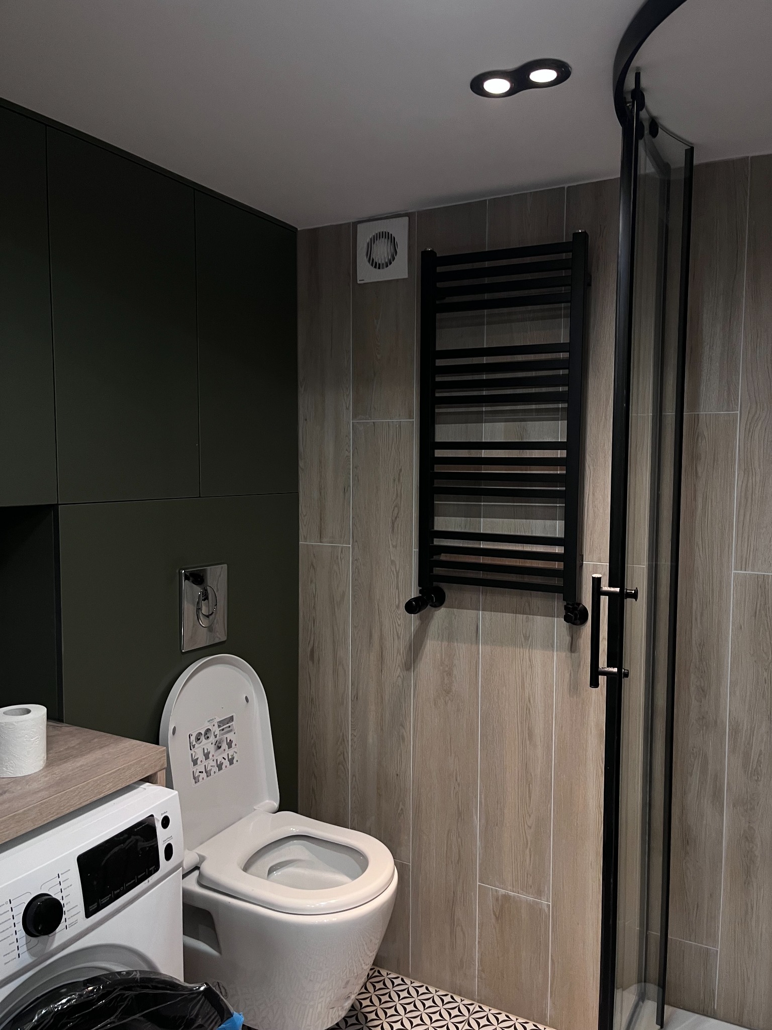 Nowoczesna łazienka z toaletą wiszącą, pralką, kabiną prysznicową z czarnymi profilami i grzejnikiem drabinkowym, wykończona płytkami imitującymi drewno i podłogą w geometryczny wzór.