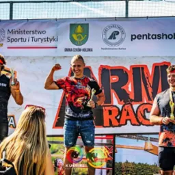 Trzej mężczyźni z trofeami na podium po biegu przeszkodowym Armagedon Race, prezentujący muskulaturę, z logotypami sponsorów w tle.