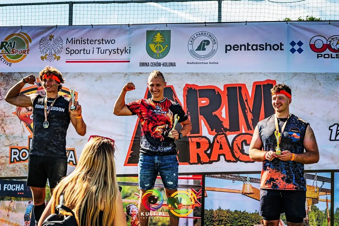 Trzej mężczyźni z trofeami na podium po biegu przeszkodowym Armagedon Race, prezentujący muskulaturę, z logotypami sponsorów w tle.