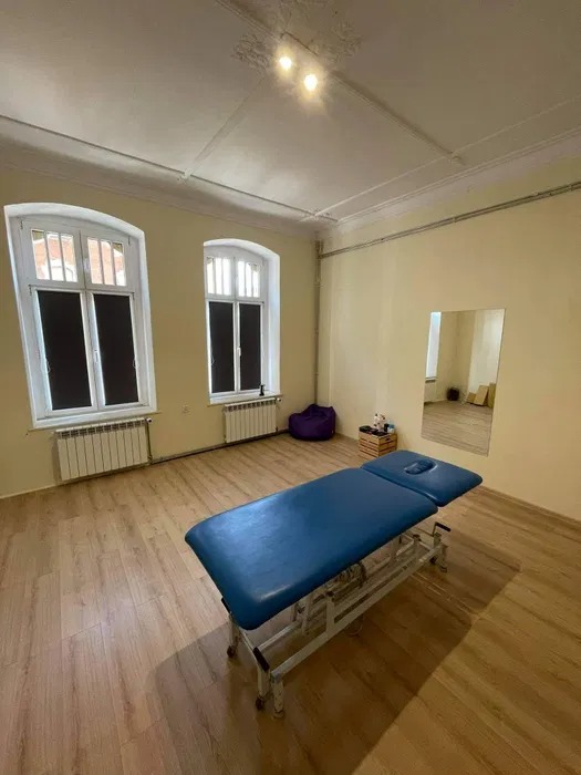 Pusty, niebieski stół do masażu w jasnym gabinecie w Katowicach, z drewnianą podłogą i dwoma oknami z żaluzjami.