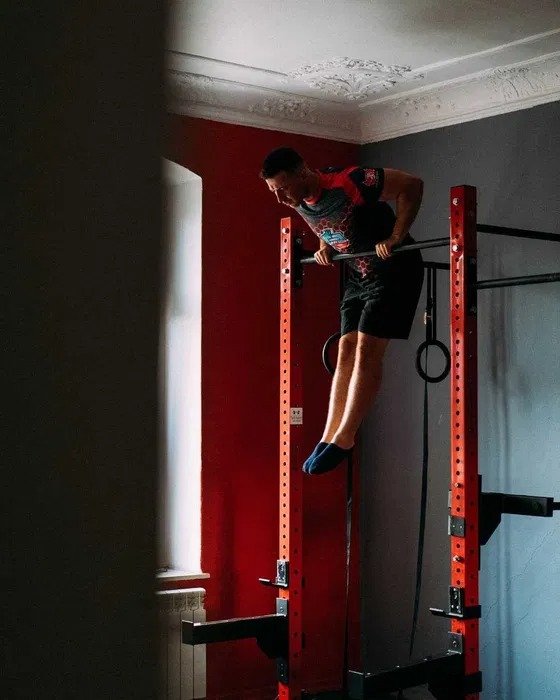 Mężczyzna wykonuje ćwiczenie na drążku w domowej siłowni z czerwonymi ścianami, w tle okno z białym grzejnikiem.