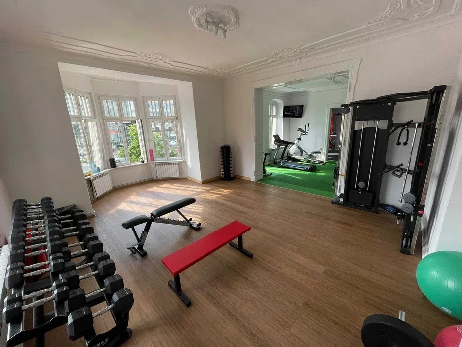 Przestronna siłownia domowa z dużym oknem, stojakiem na hantle, ławeczkami do ćwiczeń i sprzętem fitness w drugim pomieszczeniu z zieloną podłogą.