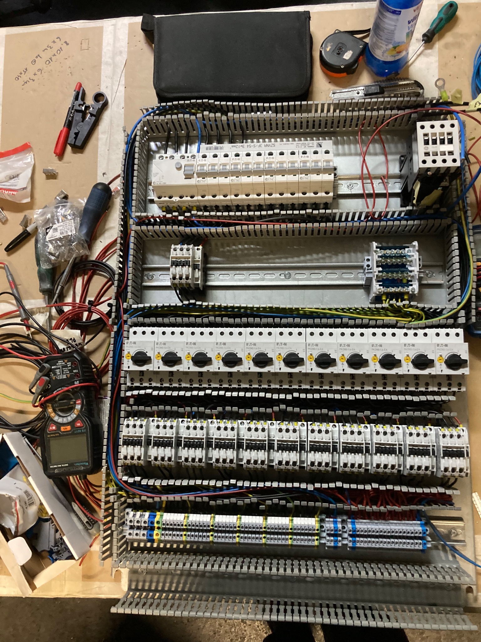 Rozdzielnica elektryczna w trakcie montażu, widoczne liczne przewody, bezpieczniki, styczniki oraz miernik elektryczny leżący obok skrzynki, narzędzia na blacie roboczym.