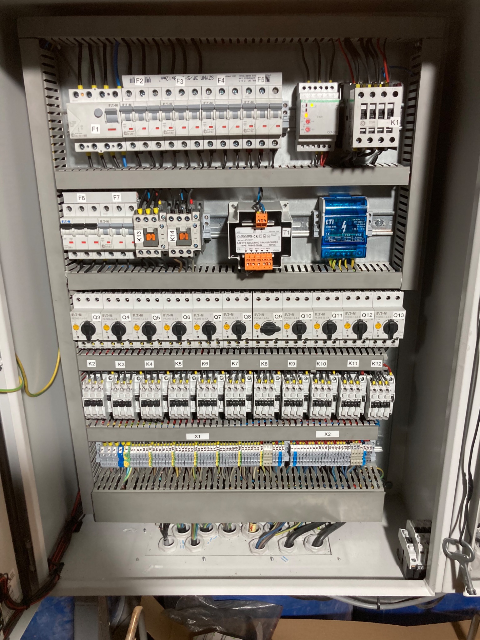 Otwarta szafa sterownicza z widocznymi elementami elektrycznymi, w tym bezpieczniki, styczniki, transformator i okablowanie, z oznaczeniami na elementach i listwach zaciskowych.