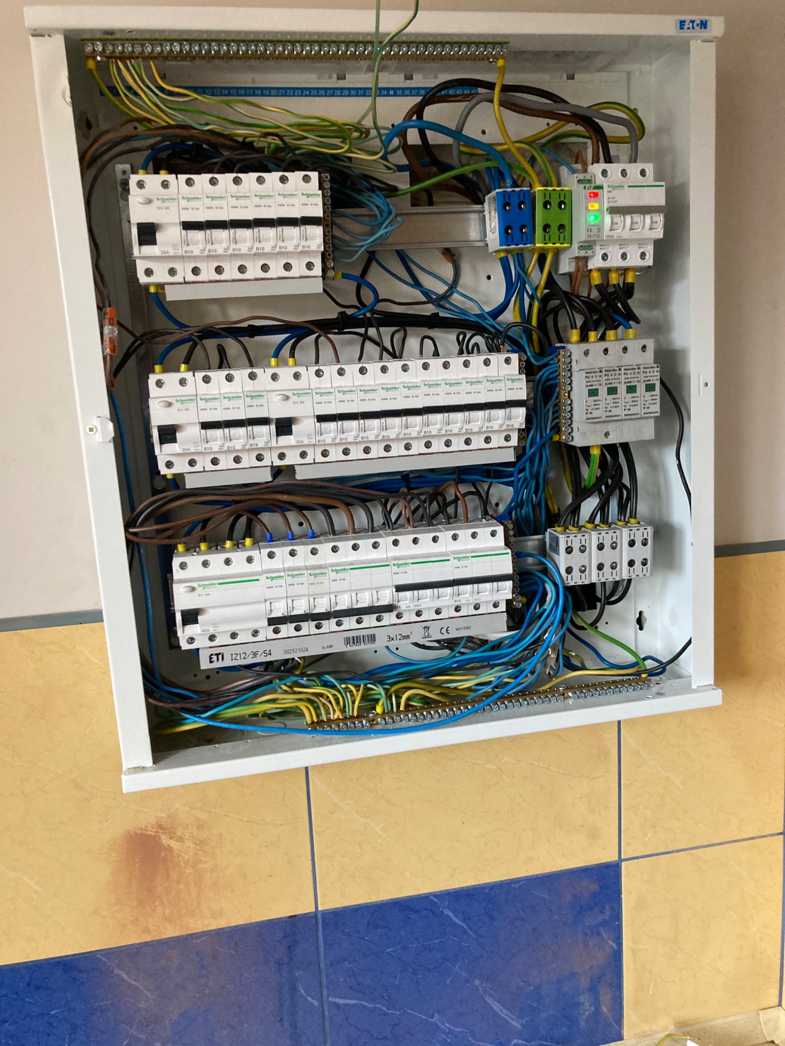 Otwarta skrzynka elektryczna z widocznymi bezpiecznikami, przewodami w różnych kolorach i elementami sterującymi; zamontowana na ścianie z żółto-niebieskimi płytkami.