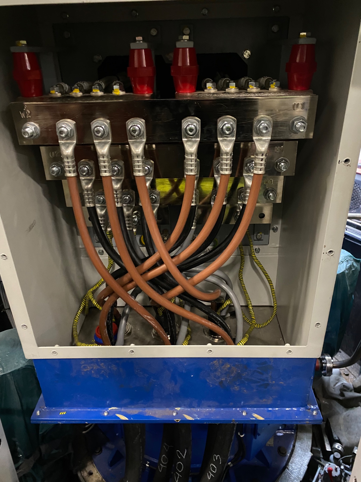 Wnętrze skrzynki elektrycznej z widocznymi przewodami w brązowej i czarnej izolacji, podłączonymi do metalowych szyn za pomocą srebrnych końcówek, z żółto-zielonym przewodem uziemiającym...