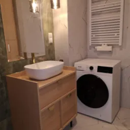 Nowoczesna łazienka z umywalką nablatową, złotą armaturą, grzejnikiem drabinkowym i pralką Gorenje na tle marmurowych płytek.