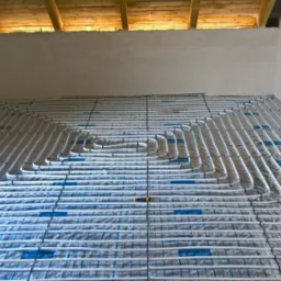 Rozłożone rury ogrzewania podłogowego na folii z siatką, widoczne elementy konstrukcyjne budynku w surowym stanie.