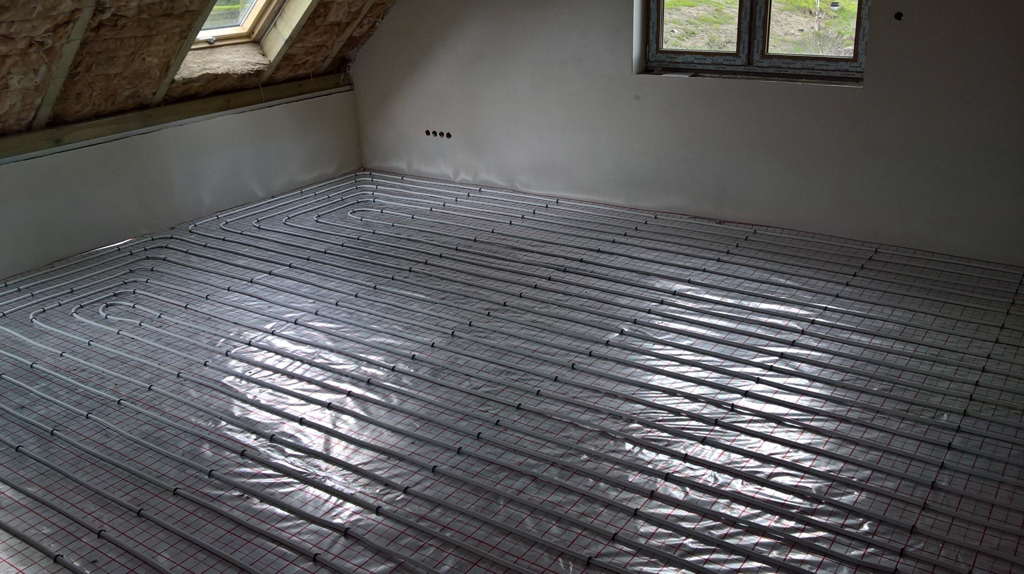 Rury ogrzewania podłogowego ułożone na folii w surowym wnętrzu poddasza z oknem dachowym i widocznymi elementami konstrukcji.