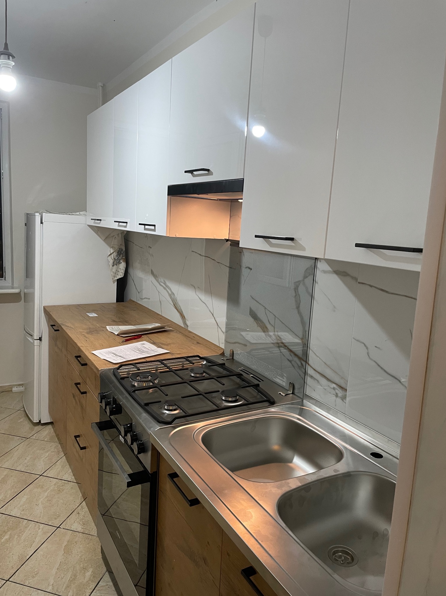 Nowoczesna kuchnia z białymi szafkami górnymi i drewnopodobnymi szafkami dolnymi, z widocznym dwukomorowym zlewem, płytą grzewczą i marmurowym panelem na ścianie.