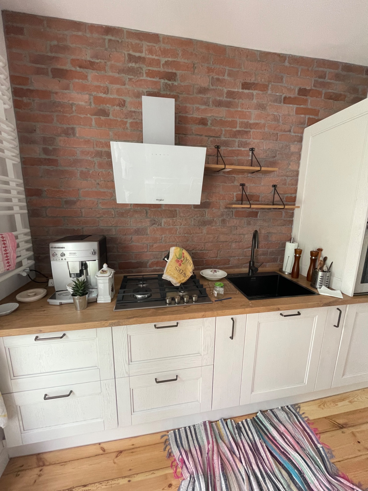 Jasna kuchnia z białymi szafkami i drewnianym blatem, ceglana ściana w tle, widoczny ekspres do kawy i czarny zlew.