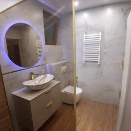 MIKOBUD Mikołaj Krauzowicz - Wykończona łazienka z okrągłym lustrem podświetlanym na niebiesko, marmurową umywalką na szafce z złotymi uchwytami i grzejnikiem drabinkowym na ścianie z płytek imitujących marmur.