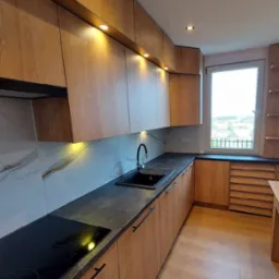 Nowoczesna kuchnia z drewnianymi szafkami, czarnym zlewem i blatem, oraz marmurowym panelem nad blatem, oświetlona punktowymi lampami sufitowymi i podszafkowymi.